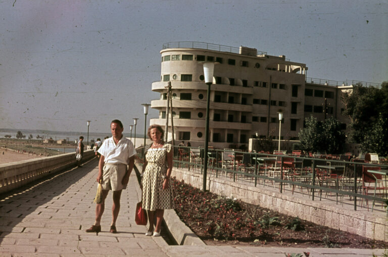 Fotografie color din 1960: un cuplu pe aleea falezei, cu Hotelul Belona și terasa lui cu scaune roșii în spate