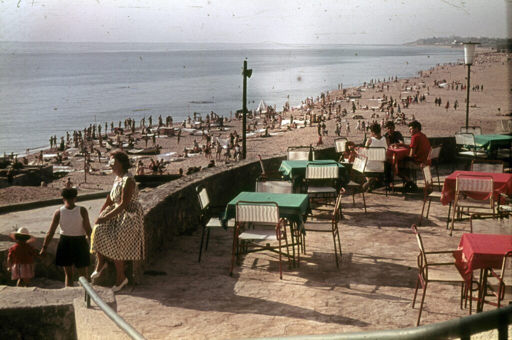 Fotografie color din 1960: terasa hotelului cu mese cu fețe de masă roșii și verzi, iar dedesubt plaja plină și marea